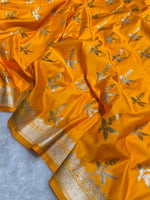 A close-up of a bright yellow-orange fabric, likely a sari, featuring metallic silver floral motifs and a wide ornate gold border.